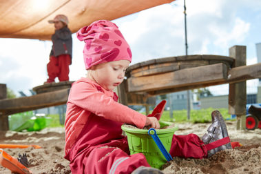 Ein kleines Mädchen sitzt unter einem Sonnensegel im Sand und spielt konzentriert mit einem Eimer und einer Schaufel..