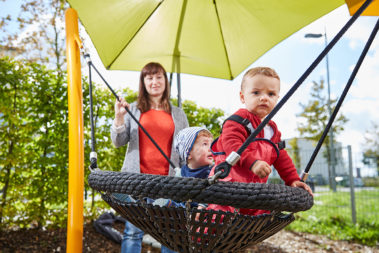 Zwei Krippenkinder schaukeln unter einem Sonnensegel und unter Aufsicht einer Erzieherin.