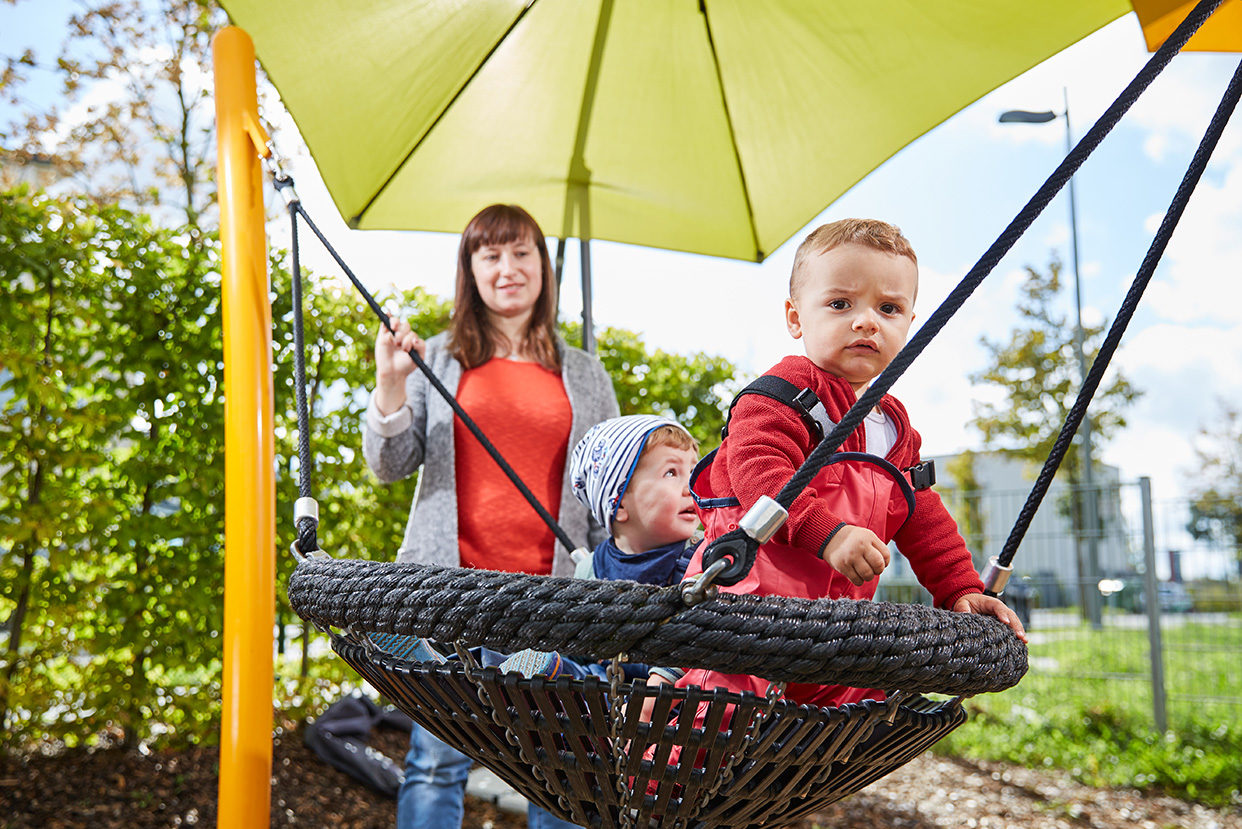Zwei Krippenkinder schaukeln unter einem Sonnensegel und unter Aufsicht einer Erzieherin.