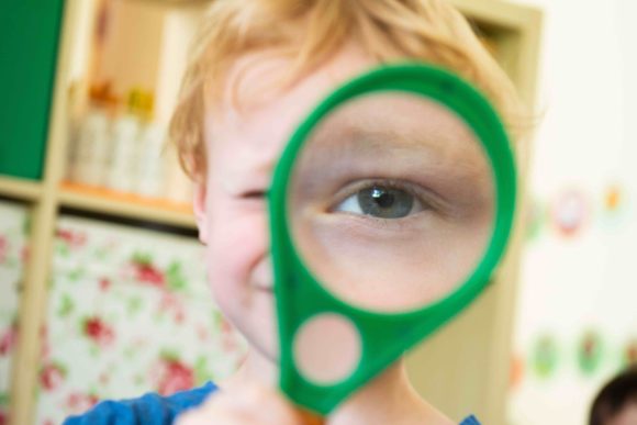 Portrait eines Kindes, das eine Lupe vors Auge hält, wodurch man das Auge im Vordergrund sehr groß sieht.