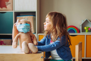 Little girl playing with rabbit soft toy in the medicine mask sitting at the table in the kids room. Ein kleines Mädchen spielt mit einem Stoffhasen, der eine medizinische Maske trägt. Sie sitzt an einem Tisch im Kinderraum.