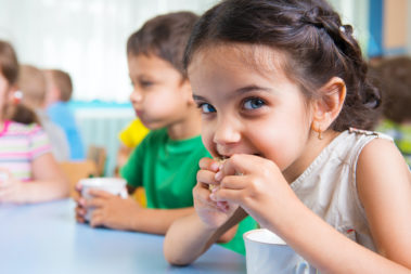 Cute little children drinking milk at daycare Ein kleines Mädchen sitzt an einem Kindergarten-Esstisch und isst einen Keks, der Junge neben ihr trinkt Milch.