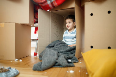 Toddler playing in cardboard box during house move, sitting in his make-believe house covered with blanket