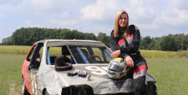 (Das Foto zeigt eine junge Frau, die in einem Rennanzug gekleidet und gestützt auf einen Remmhalm auf der Motorhaube eines ramponierten Autos sitzt. Davor steht ein kleines, rotes Rutschauto (Bobbycar).) Das Foto zeigt eine junge Frau, die in einem Rennanzug gekleidet und gestützt auf einen Remmhalm auf der Motorhaube eines ramponierten Autos sitzt. Davor steht ein kleines, rotes Rutschauto (Bobbycar).