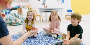 Kindergartenkinder sitzen im Halbkreis auf einem Spielteppich, vor ihnen eine Erzieherin (Kindergartenkinder sitzen im Halbkreis auf einem Spielteppich, vor ihnen eine Erzieherin) Kindergartenkinder sitzen im Halbkreis auf einem Spielteppich, vor ihnen eine Erzieherin