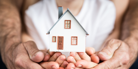 Kinderhände, gestützt von Erwachsenenhänden, halten das Modell von einem kleinen Haus.